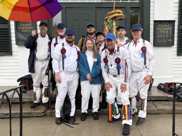 Nine members of Newtowne Morris standing on a on the steps of a church wearing whites, bells, suspenders, and blue caps, with a rainbow umbrella on the back left and the ki lin to the back right.